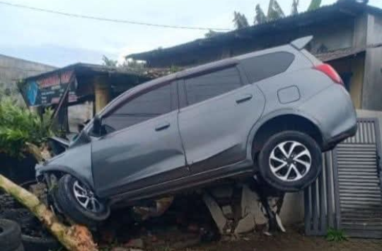Mobil minibus berwarna abu-abu tampak tersangkut di teras rumah warga setelah menabrak pohon di Desa Perdamaian, Kecamatan Binjai, Langkat, Minggu (16/11/2025).